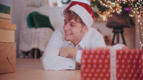 Un-Hombre-Alegre-Con-Sombrero-De-Papá-Noel-Deslizando-Regalos-De-Navidad-Mientras-Está-Acostado-En-El-Suelo