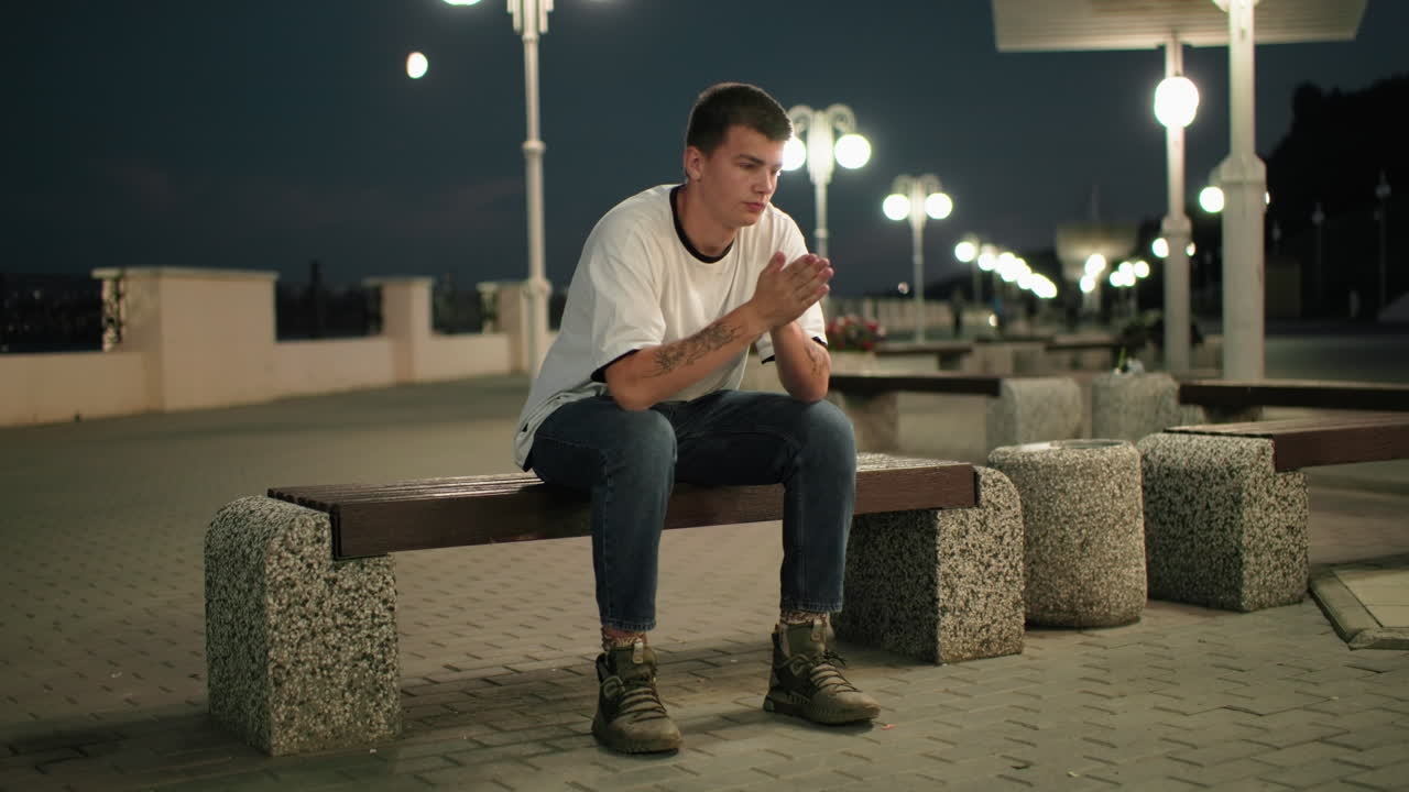 joven adulto sentado en un banco al aire libre por la noche, manos en la cara y luego quitando la mano, mirando ansioso o estresado, luces de la ciudad y peatones borrosos en el fondo