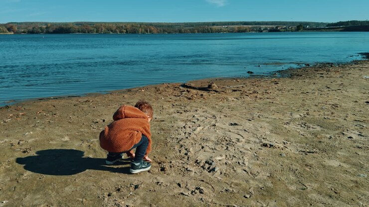 Caucasian boy touching sand on the river bank. Cute kid takes some handful of sand and throws it into water.