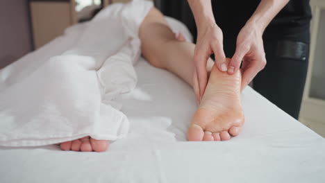 massage therapist using both thumbs to apply gentle pressure on client sole and side foot during wellness treatment, showing clean environment with white linens and relaxed body posture