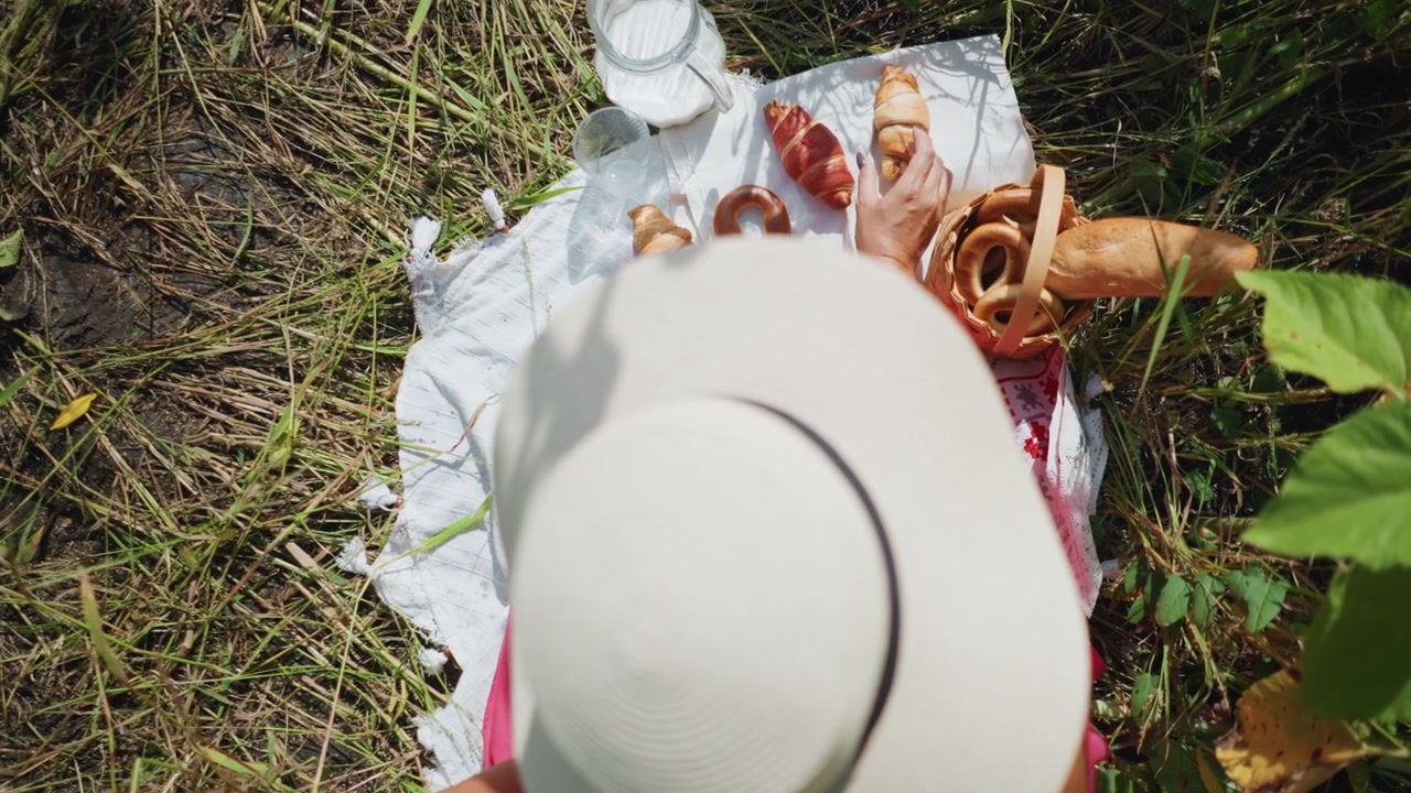 Een vrouw met een witte hoed geniet van het kiezen van gebak van een buitenpicknick op het gras, inclusief croissants en baguettes. Een geweven mand bevat broodjes naast een deken met glazen en een melkkan.