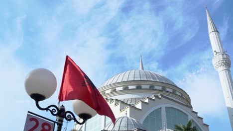 flag of albania flutters in the wind on warm sunny day, blue cloudy sky and beautiful white stone mosque in background. national attributes, monument of architecture