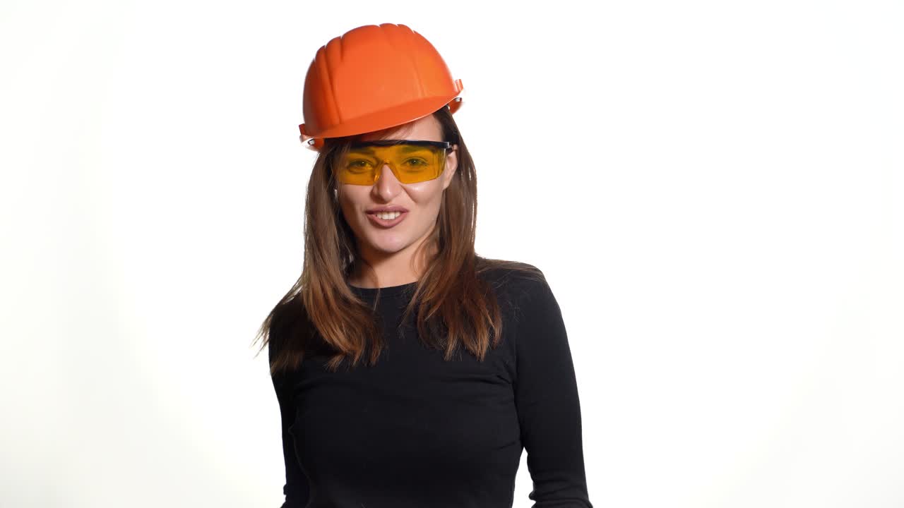 Joyful woman engineer in spectacles is standing in an orange helmet and posing on a white background.