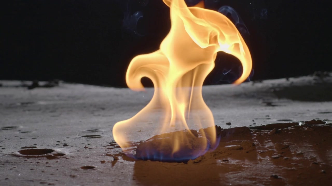 Eine kleine Kraftstoffpfütze entzündet sich und erzeugt eine sich kräuselnde gelbe Flamme auf einer schwach beleuchteten Tischplatte, mit Tropfen und Rauch.