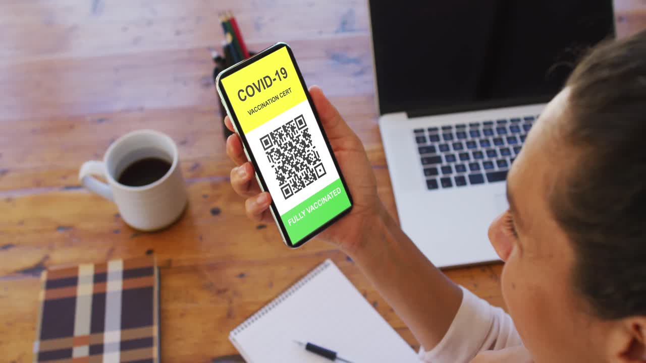 Woman at desk holding smartphone with covid vaccination certificate and qr code on screen