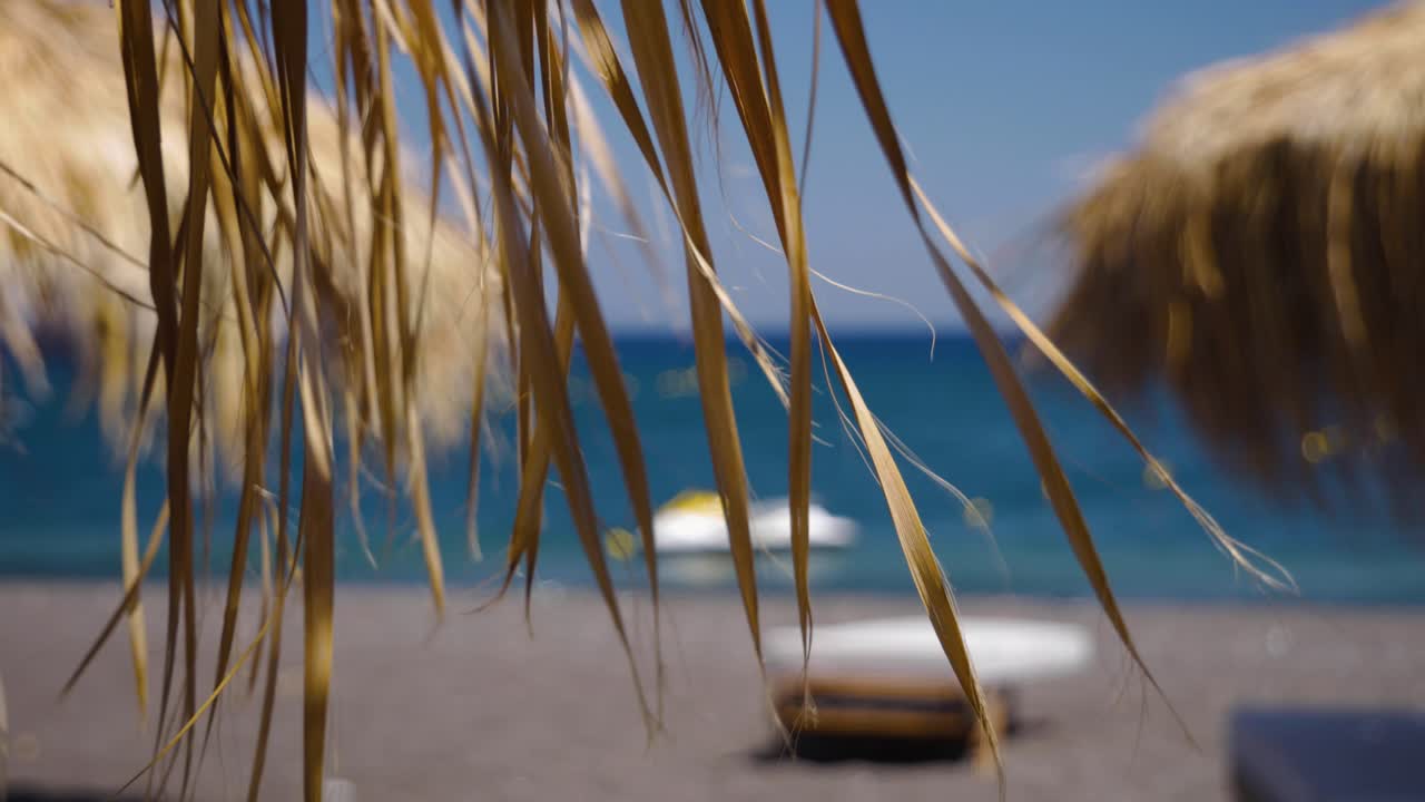 Primo piano a mano libera di foglie di palma secche mosse dal vento da un ombrellone di paglia sulla spiaggia di lava nera a Perissa, Santorini, in Grecia, Europa.