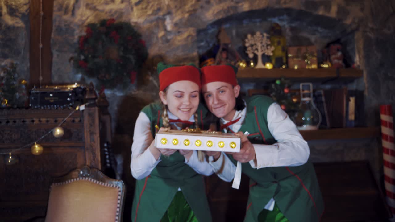 Joyful elves with magic book in decorated room at Christmas. Two elves in green costumes holding beautiful box and blowing out sparkles from top.