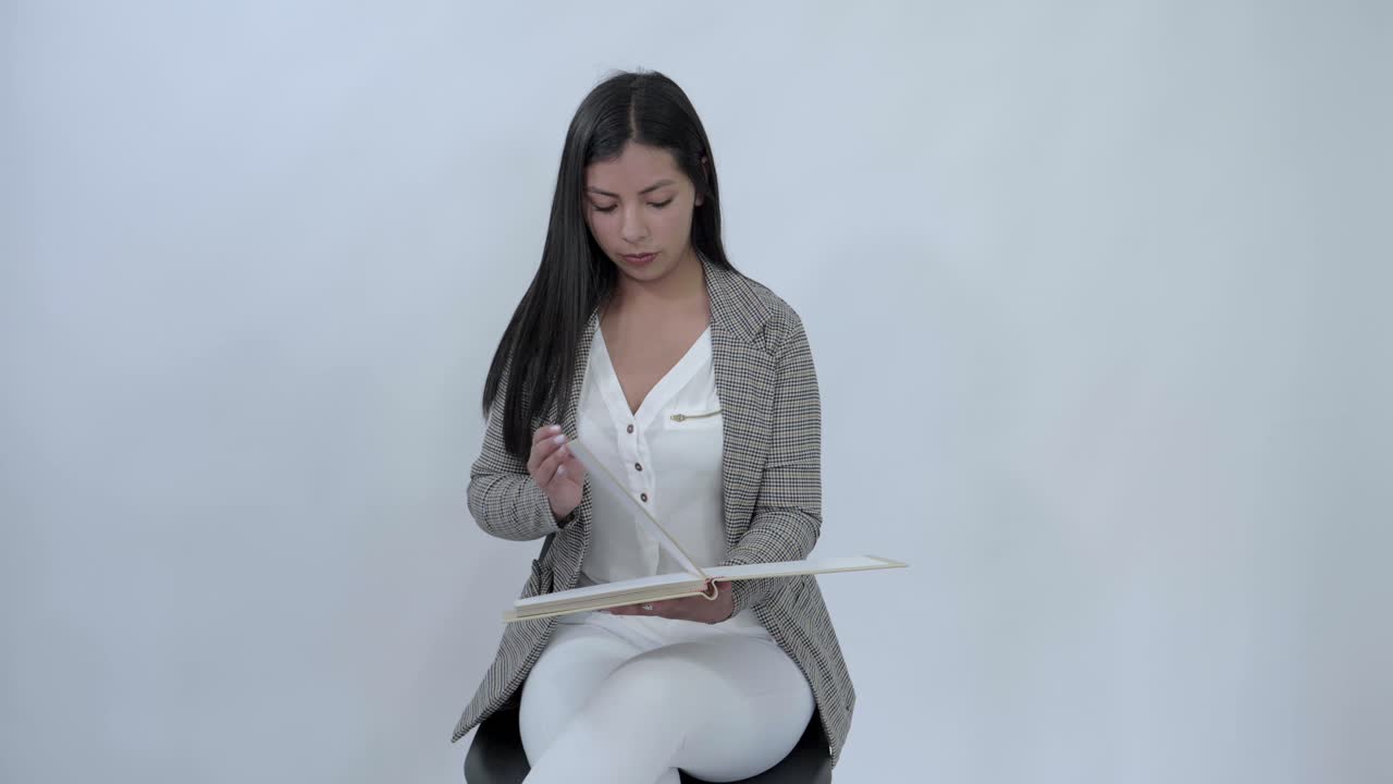 Latina girl sitting in a chair reviews a book, turning each page one by one, against a white backdrop, displaying concentration in her quiet study