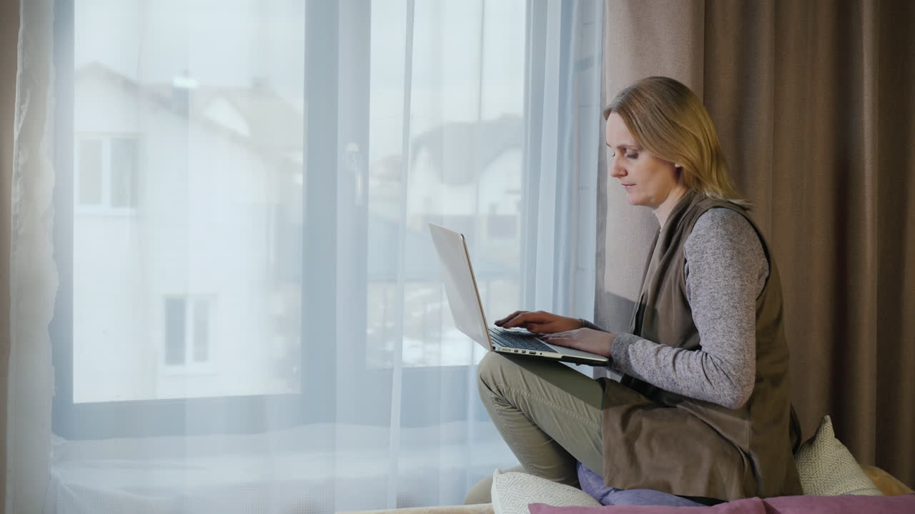 A woman sits on the window sill in her room and uses a laptop