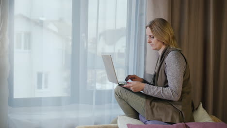 una mujer se sienta en el alféizar de la ventana de su habitación y usa una computadora portátil
