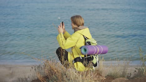 Junger-Mann-Mit-Rucksack-Fotografiert-Das-Meer-Auf-Dem-Smartphone,-Während-Er-An-Der-Felsigen-Küste-Entlang-Spaziert