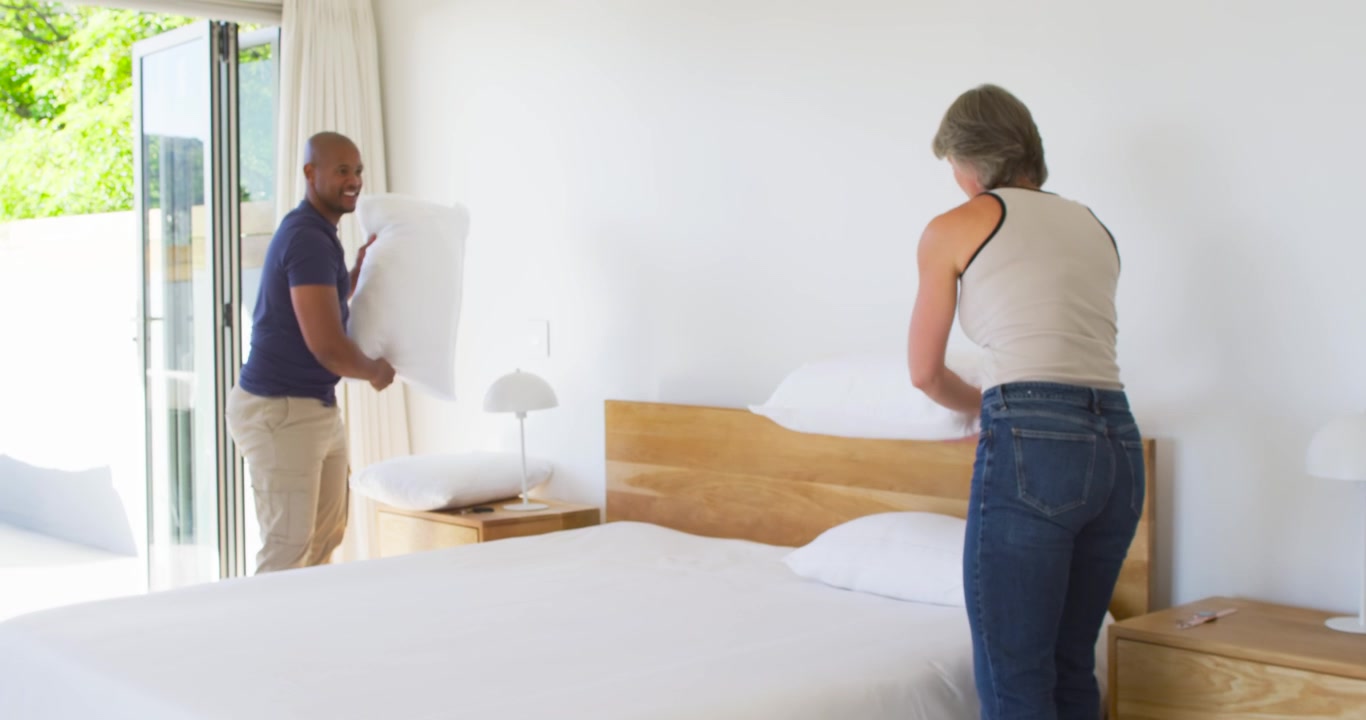Couple diversifié arrangeant et empilant des oreillers blancs pour finir de faire un lit king-size, lissant la couette.
