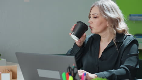 woman takes short break to sip latte while working on laptop at office desk surrounded by building model, organized documents, green plant, and writing tools in modern business environment