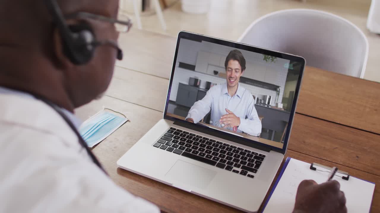 Ein afroamerikanischer Arzt mit Telefon-Headset macht sich Notizen während eines Videoanrufs am Laptop.