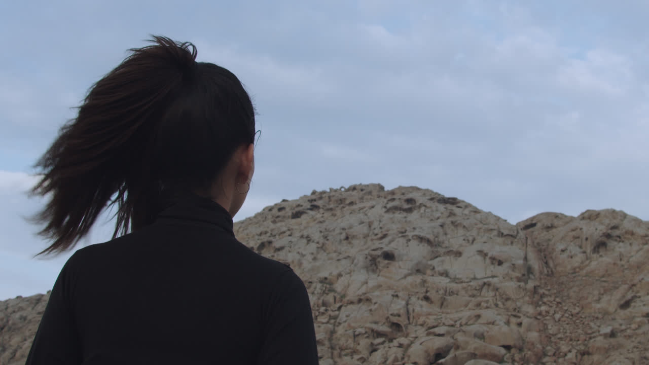 primer plano en cámara lenta de una mujer misteriosa vestida de negro en el desierto mirando montañas de arena, mientras el viento mueve su pelo largo