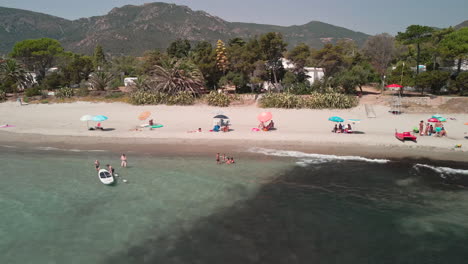 Tourist-Of-Families-Under-Their-Big-Umbrellas-Enjoying-Summer-Holidays-On-Sandy-Shoreline-At-Sardinia,-Island-Of-Italy