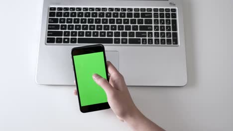 male hands scrolling on a larger black smartphone, laptpo in the background. green screen, chromakey concept, laptop in the background. top shot 60 fps