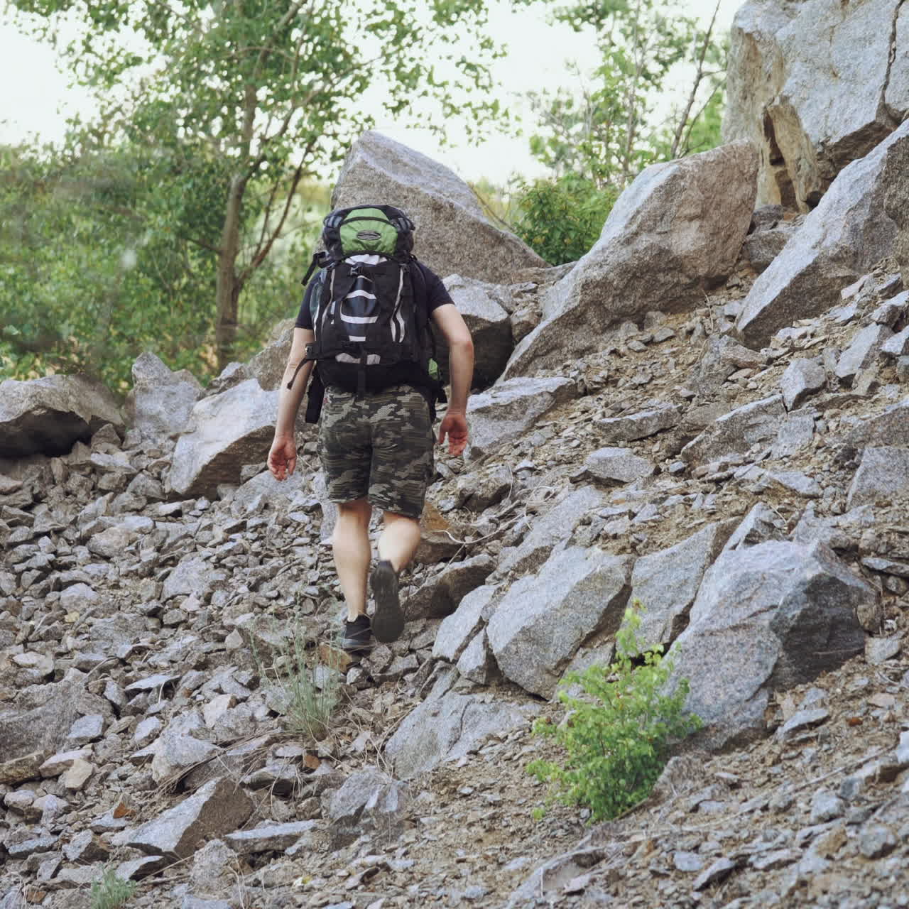 A traveler with a backpack on his shoulders rises to the top of the mountain with stones in the afternoon in warm weather. Tourism.