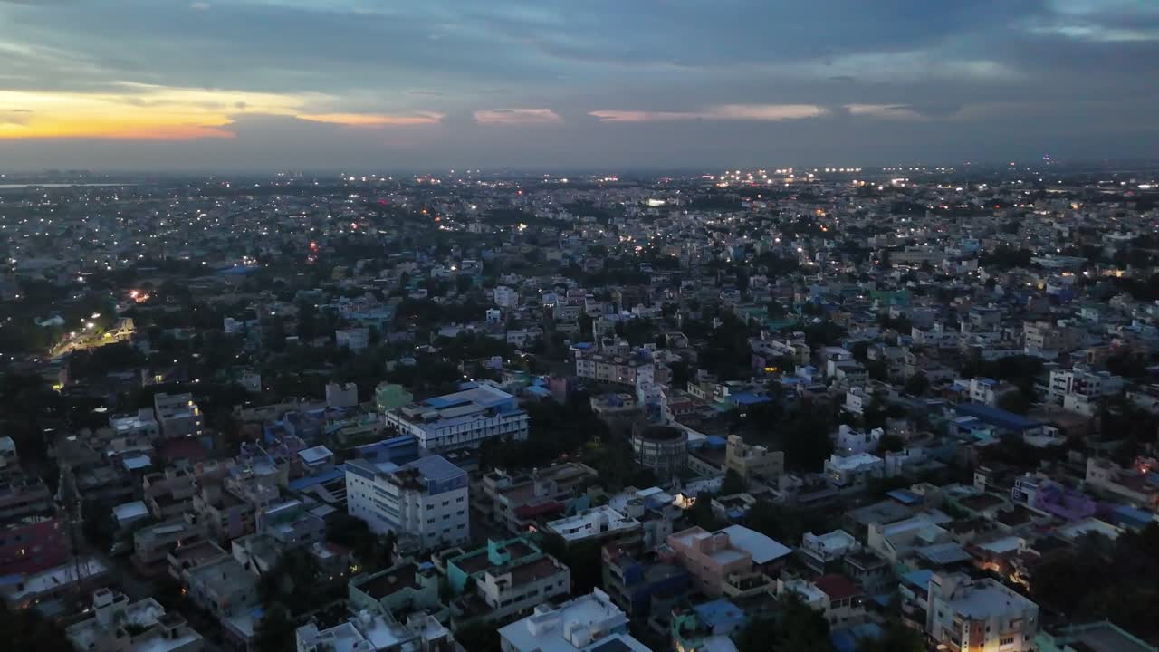 A wide aerial shot of Chennai at dusk, contrasting older low-rise neighborhoods with modern high-rise apartments. Streetlights and window glows animate the city as faint hills line the distant horizon