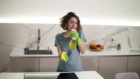 Una-Linda-Mujer-De-Pelo-Corto-Con-Guantes-Protectores-Verdes-Canta-Usando-Un-Aerosol-Químico-Como-Micrófono-Mientras-Limpia-La-Superficie-De-La-Estufa-De-Cerámica-Con-Un-Trapeador.-Canto-Emocional-Durante-La-Limpieza