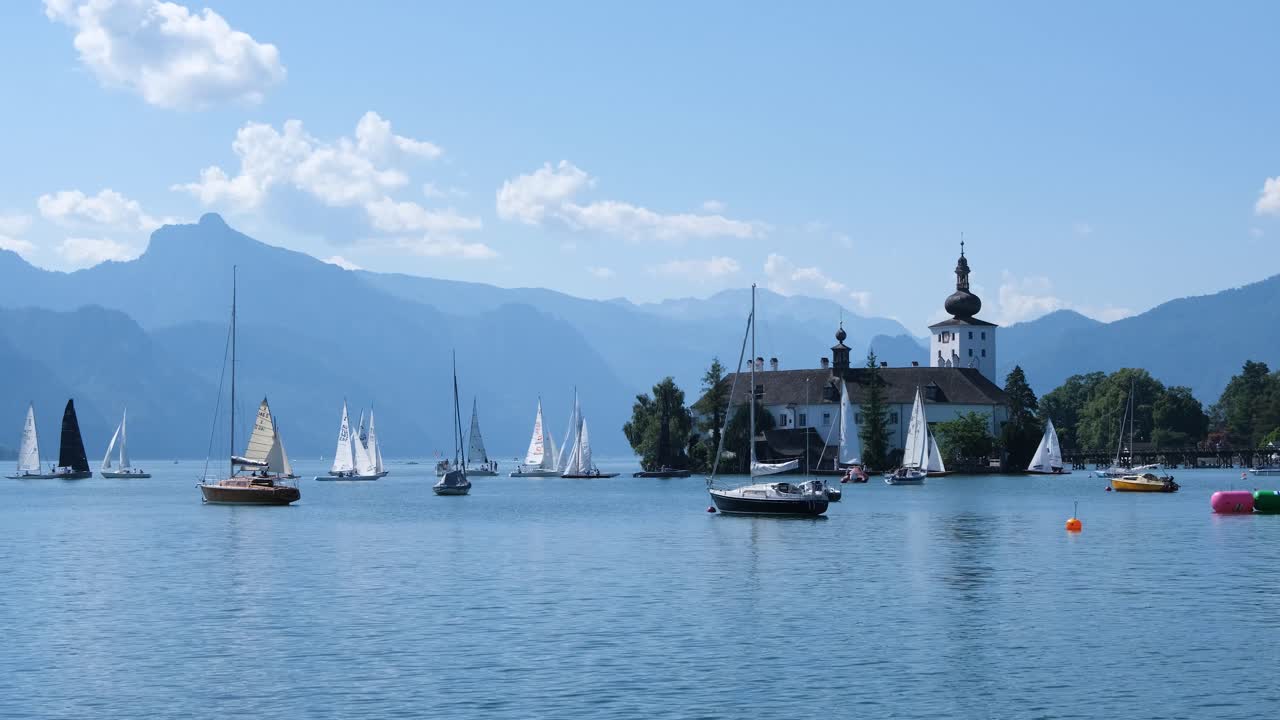 Serene footage of a calm lake dotted with colorful sailboats, some gliding gently while others rest with sails down. In the background. majestic mountains rise beneath a clear blue sky