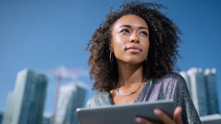 A Thoughtful Woman with Curly Hair Holding a Tablet Gazes into the Distance Against a Bright Blue Sky and Urban Background, Reflecting Determination and Vision for the Future