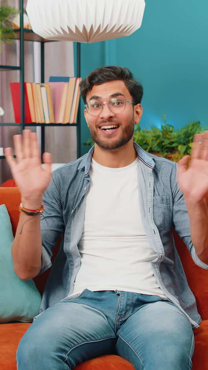 hombre indio sonriendo amistosamente a la cámara agitando las manos gestando hola hola saludando en casa en el sofá