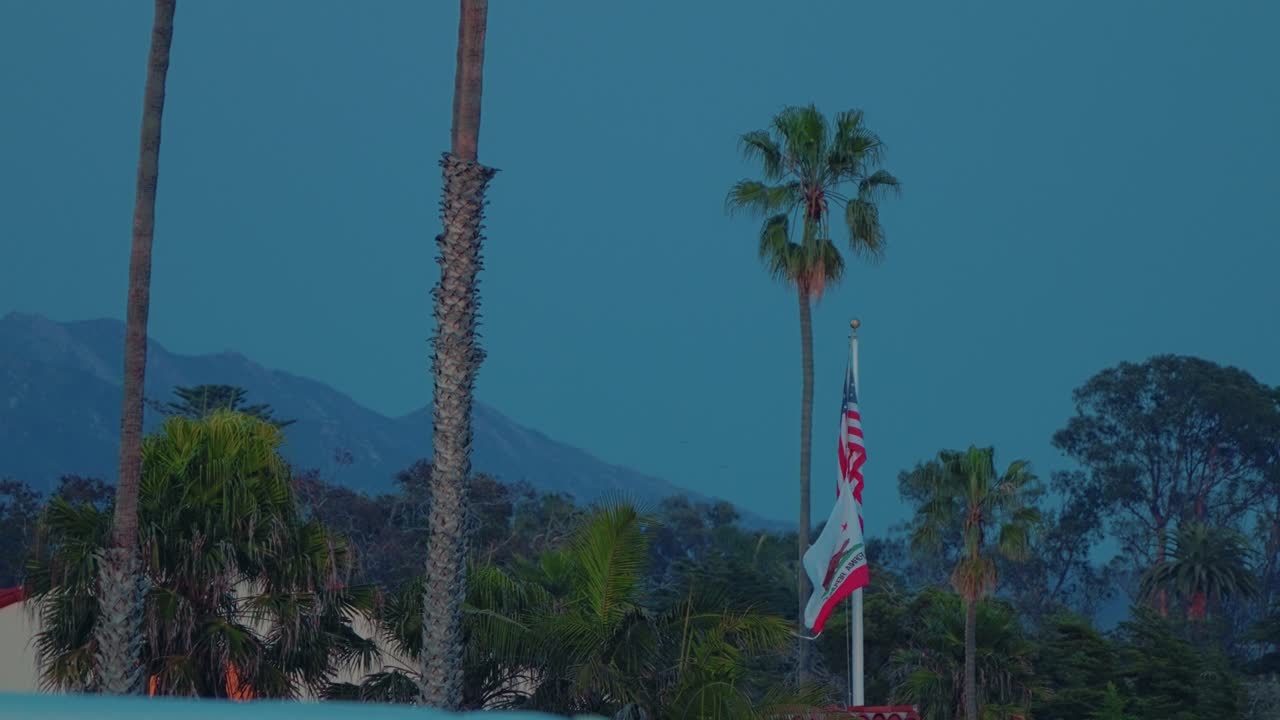 American and California Flags Waving at Twilight with Palm Trees and Mountain Backdrop in Santa Barbara
