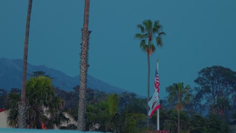 american and california flags waving at twilight with palm trees and mountain backdrop in santa barbara
