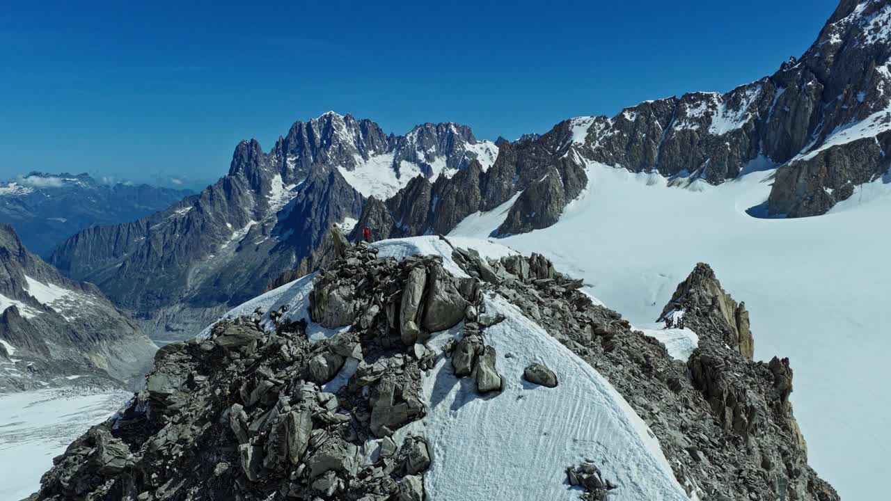 Chamonix yakınlarındaki Fransız Alpleri'nde, berrak mavi gökyüzü altında devasa sivri zirveler ve buzullarla çevrili, engebeli karlı bir dağ sırtında ilerleyen bir tırmanıcının geniş açılı havadan görünümü.