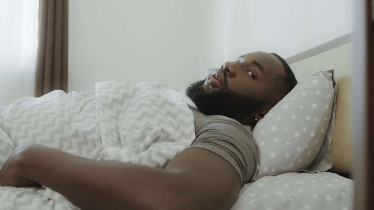 Uomo di colore scioccato seduto a letto al mattino. Giovane adulto che prende il telefono a letto.