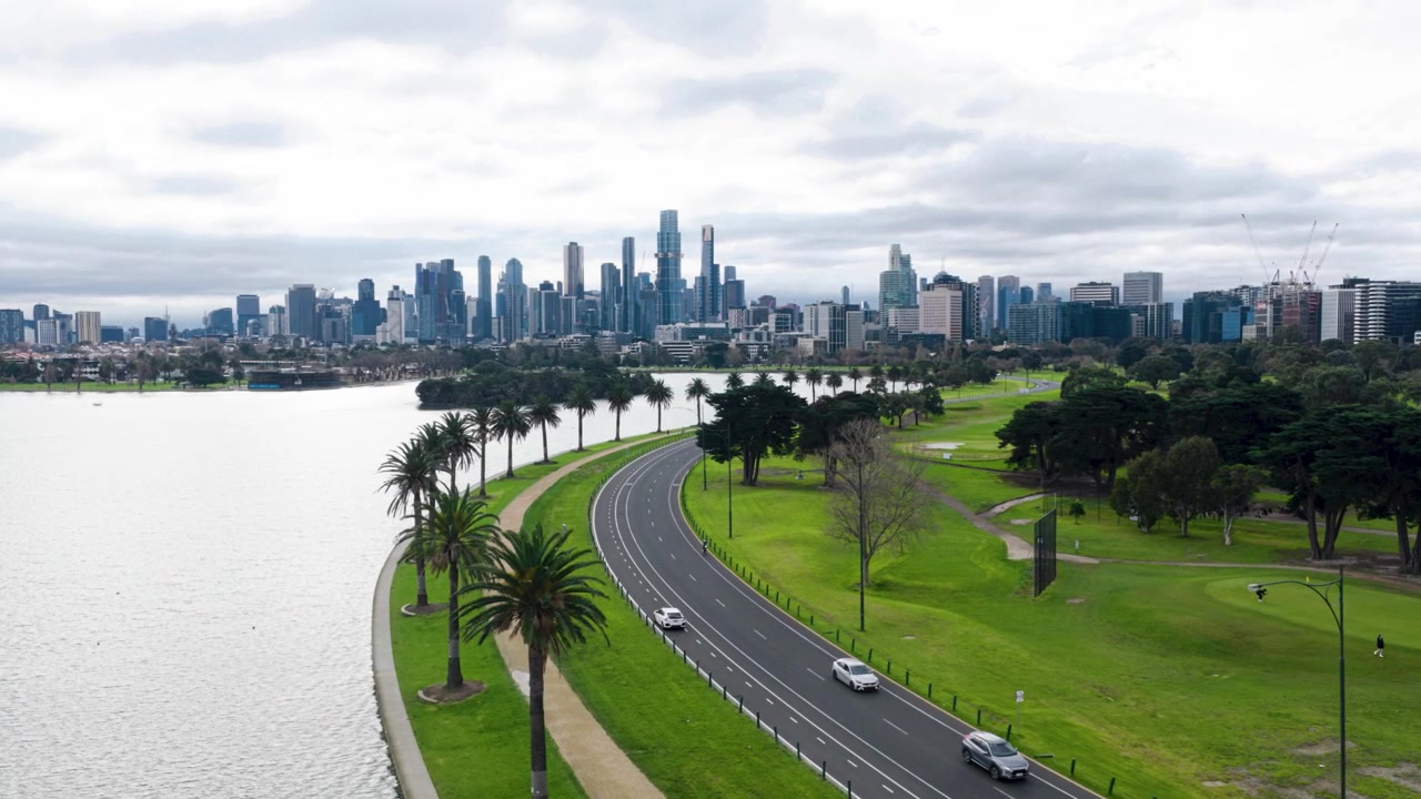 Splendide riprese aeree cinematografiche con drone del lago Albert Park che mostrano la strada curva lungo il lago con traffico in movimento, ciclisti, palme, ampi parchi e lo skyline del CBD di Melbourne come sfondo.