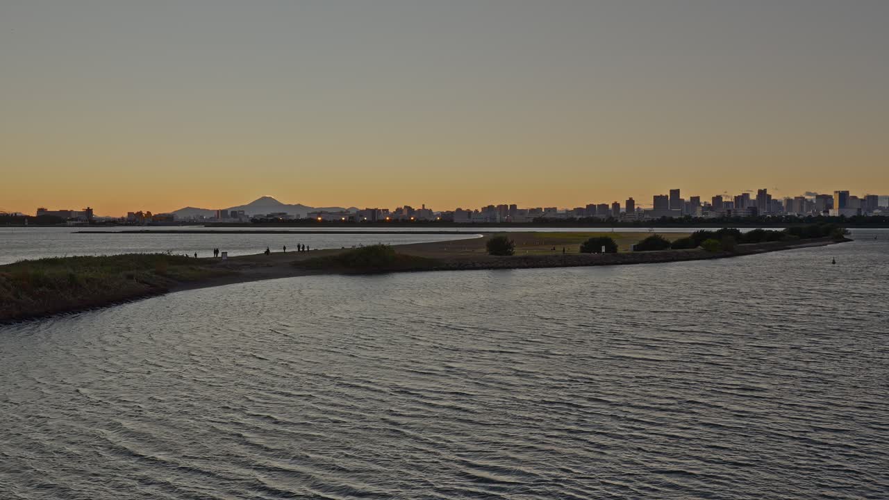 A low-angle shot over the water showing a long spit of land, the Tokyo skyline, and Mount Fuji's peak at sunset