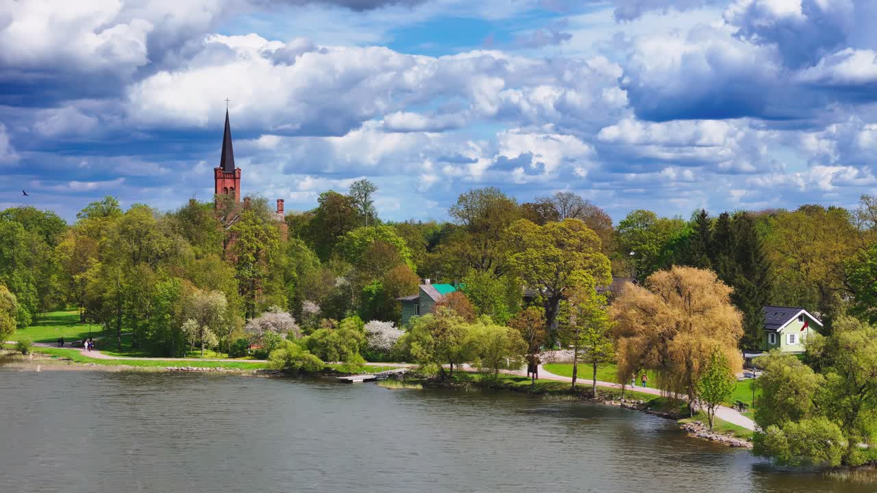idyllic spring day in Vaxjo, Sweden, captured from an elevated wide-angle view, showcasing vibrant green trees, a distant red-brick church spire, and fluffy white clouds reflected in the calm lak