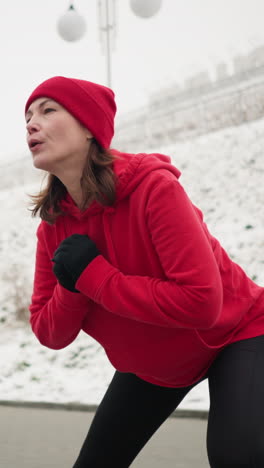 coach working out in winter, stretching into focused side lunge on paved ground, arms clamped close to chest, wearing hoodie and black leggings, exercising near snowy hill with urban surroundings