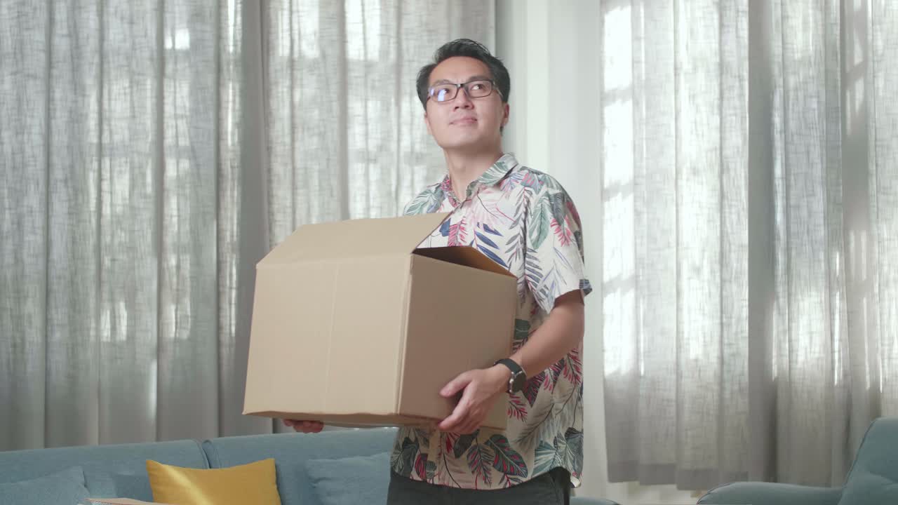 hombre asiático llevando una caja de cartón con cosas en una casa nueva y mirando a su alrededor. se muda a una casa nueva