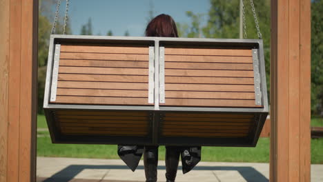 back view of lady on wooden swing chair with hair fluttering in wind, swinging gently in serene park surrounded by lush greenery and trees under bright sky