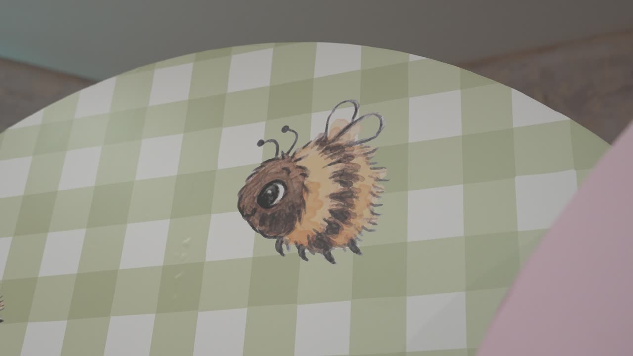 En person visar upp ett målat bi på ett runt bord med ett grönt rutigt mönster. Biet är färgglatt och sticker ut mot bakgrundsdesignen