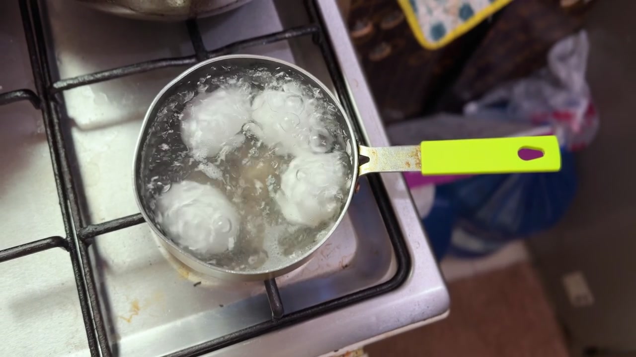 Quattro uova bianche bollono in una piccola pentola di metallo su un fornello a gas. L'acqua che bolle vigorosamente copre le uova durante la preparazione di una colazione sana in una cucina di casa.
