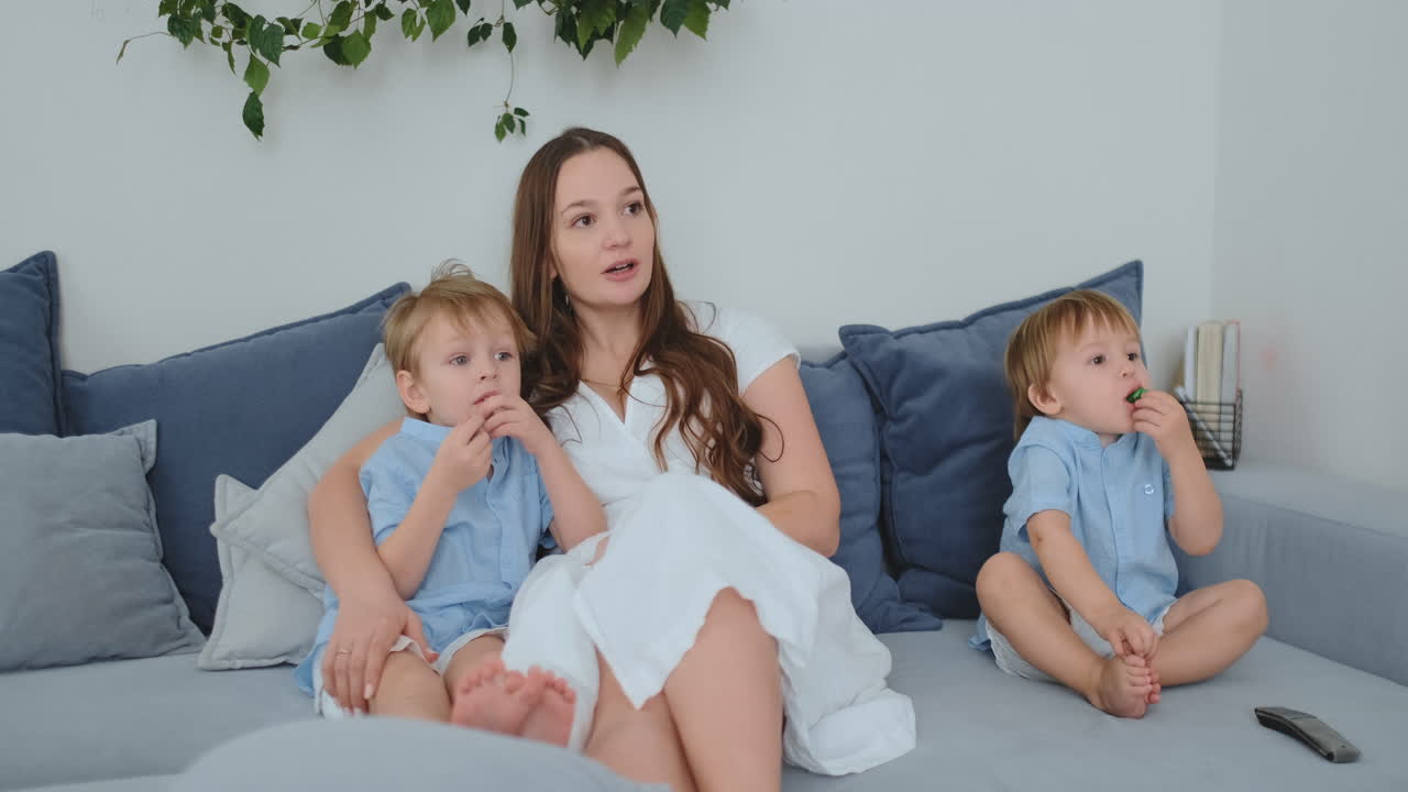 엄마는 거실의 소파에서 두 작은 아들을 안아 tv를 보고 있습니다. 가족은 tv를 보고 있습니다.