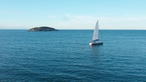 small sailboat sails at joao fernandes beach, rio de janeiro brazil, tropical blue water and green lonely island in the ocean, aerial drone shot