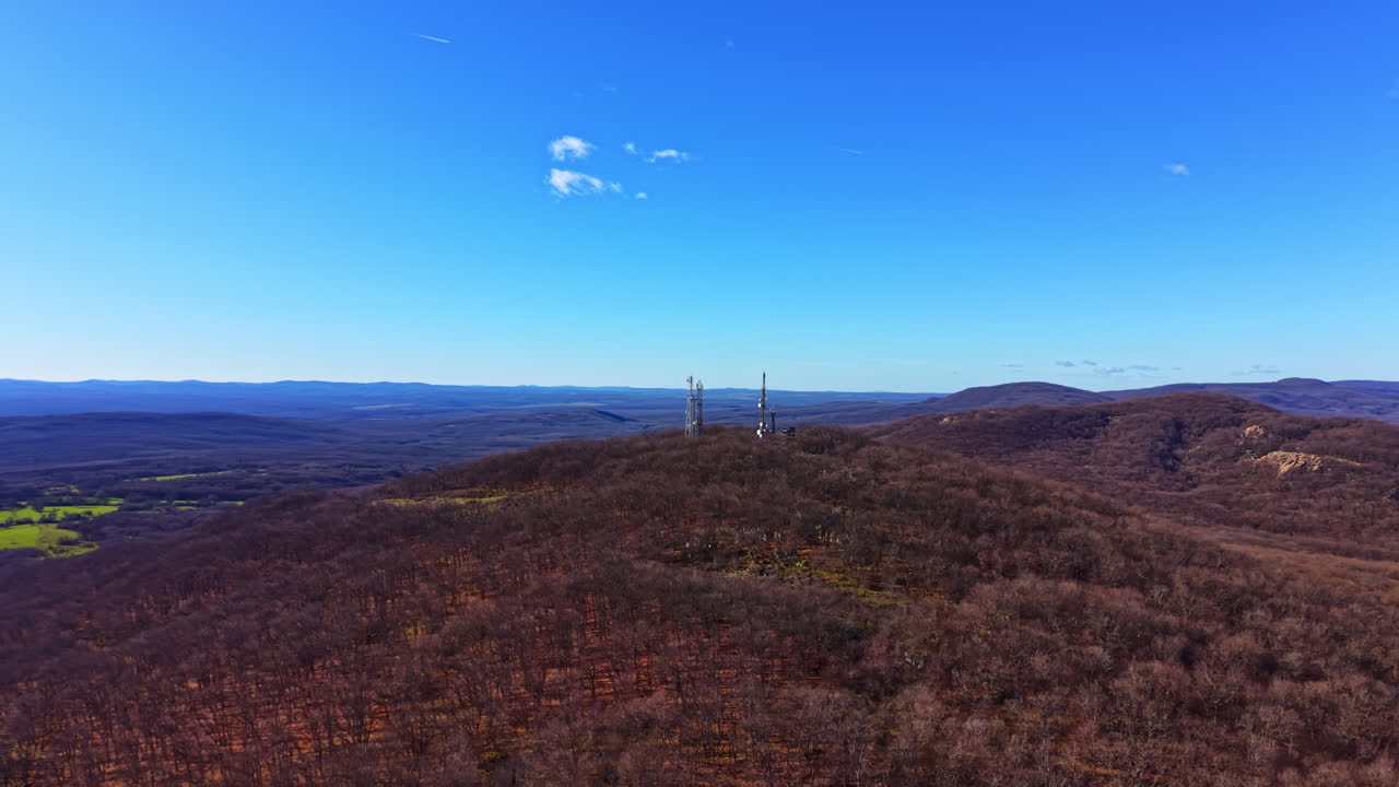 Mountaintop view shows communication towers in a landscape with trees and hills under a clear blue sky during daytime