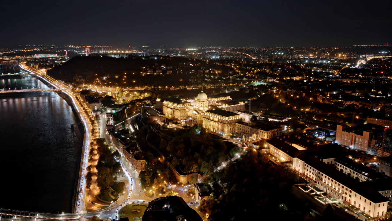 A glowing night view of Buda Castle perched above the Danube, surrounded by twinkling city lights and winding riverside roads