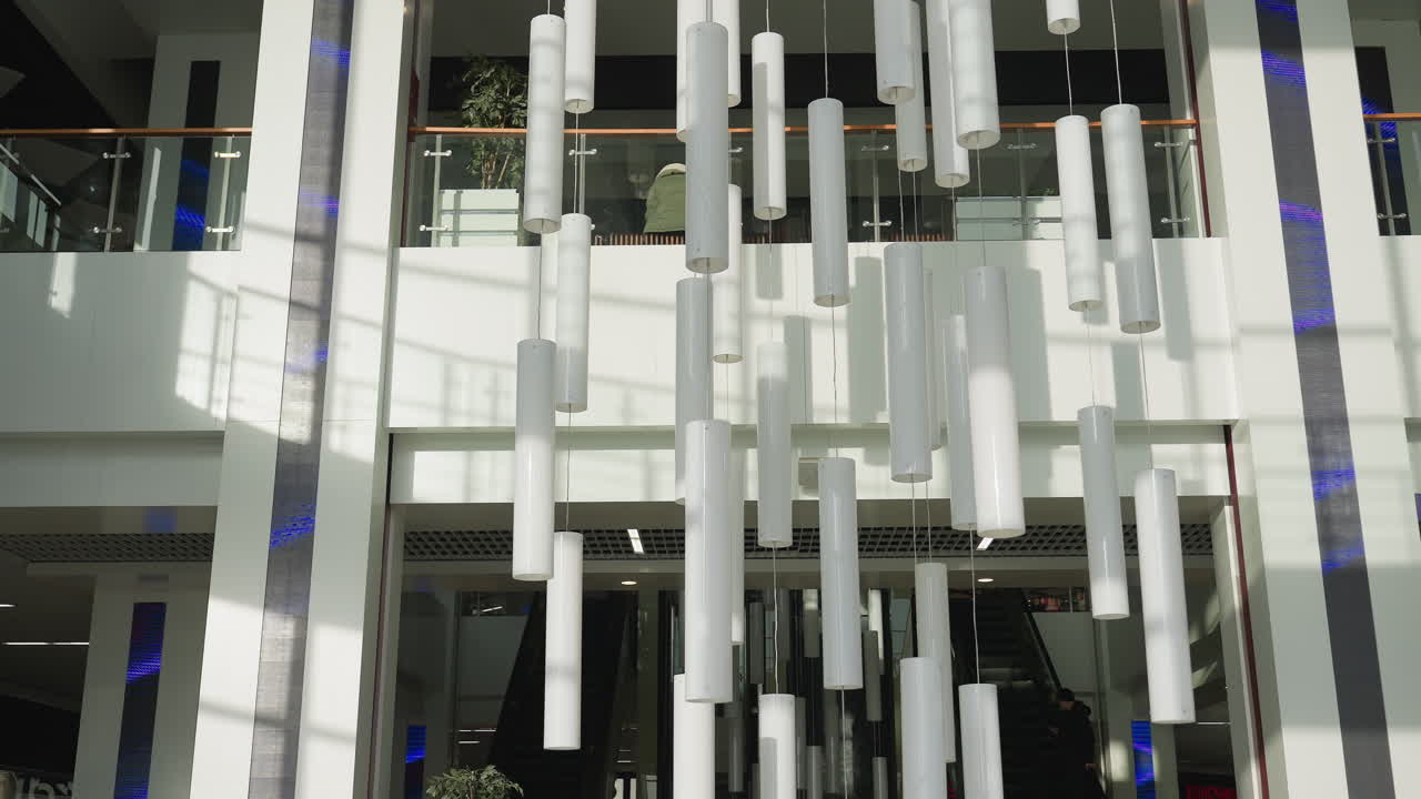 Interior of stylish shopping mall featuring contemporary cylindrical hanging lights, glass railings, and escalator centered between walls, with clean architectural lines