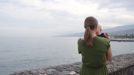 Walking-by-the-ocean-on-an-ancient-European-square,-a-young-mother-enjoys-the-company-of-her-baby-son,-gazing-at-the-waves-and-exchanging-smiles