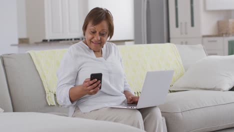 Feliz-Mujer-Mayor-Afroamericana-Sentada-En-El-Sofá-Usando-Un-Teléfono-Inteligente-Y-Una-Computadora-Portátil,-Sonriendo