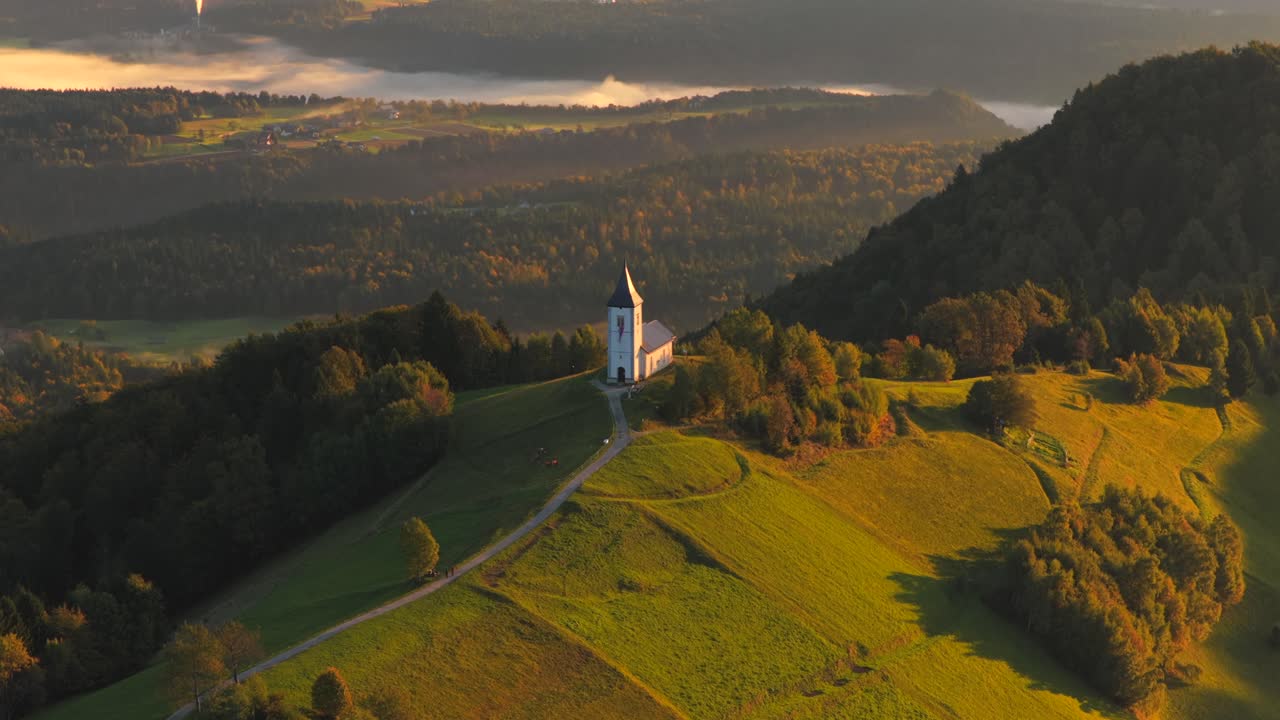 Aerial paralax shot of famous Jamnik church during golden sunset in Slovenia. Sava river under fog in the valley. Wide shot