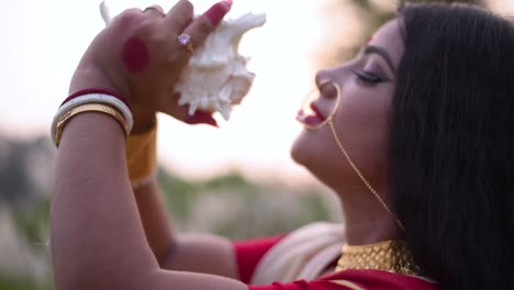 A-Hindu-married-Indian-woman-is-about-to-blow-a-conch-shell-or-shankha-in-a-relgious-way-in-a-field-at-sunset-or-sunrise,-gold-jewellery