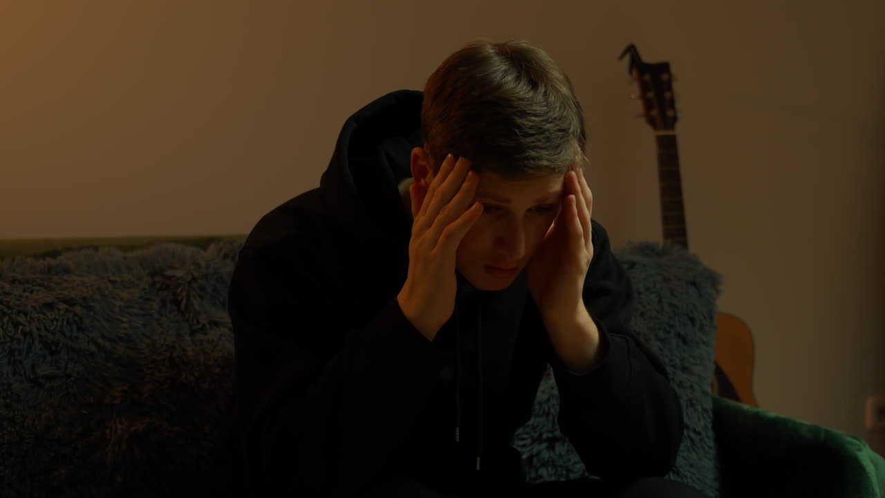 Young caucasian man sitting on a couch with acoutic guitar standing near, holding his head with hands, looking down in distress, symbolizing mental health issues such as stress, anxiety and fatigue