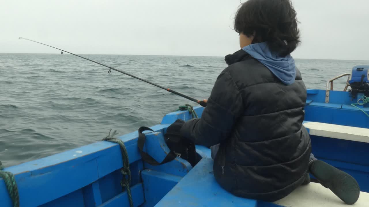 Seorang anak laki-laki sedang memancing di perahu biru. Anak itu mengayunkan kembali pancingnya dan melempar. Perahu bergoyang dan bergerak di lautan biru luas yang membentang hingga ke cakrawala